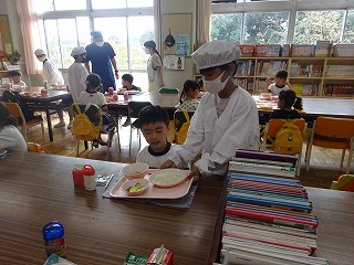 白い給食着とマスク、三角巾を着用した児童が、園児のテーブルまで給食が乗ったトレイを運んでいる様子の写真