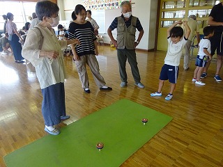 広い室内の床に敷かれた緑色のマットの上で2つの駒が回り、男の子が両手を上げ喜び、それを取り囲むように大人が見守っている写真