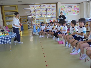 壁に掲示物が飾られた室内で、一人の女性が子供たちの前でハンドベルのような楽器を鳴らしており、子供たちが横一列に椅子に座ってハンドベルを持ち練習している写真