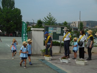 門の前で青いスモックと帽子をかぶった園児たちに、挨拶運動のたすきをかけた先生や地域の人々が園児に挨拶をしている写真