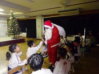 飾り付けされたクリスマスツリーが置かれた室内で、サンタクロースに扮した大人が集まった園児たちとハイタッチをして交流している様子の写真