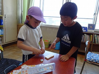 紫色の帽子をかぶった体操服姿の園児が黒いティシャツを着た児童に机で四角いブロックのようなものに丸いシールを張るのを教わっているような写真