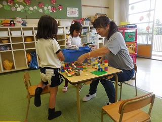 広々とした教室で二人の園児がテーブルを囲み、座って一緒に遊ぶ先生とカラフルなブロックを使って楽しそうに遊ぶ様子を捉えた写真