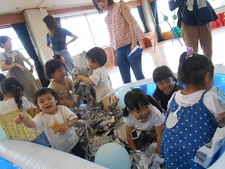 教室で大きなビニールプールの中で、破いた新聞紙を持ち上げ数人の子供たちが満面の笑みで遊んでいる様子をプールの外から数人の保護者が見守っている写真