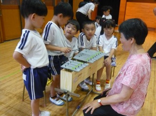 体育館のような室内で、体操服姿の五人の園児たちが木製の台に置かれた鉄琴を囲み、座って見守っている先生の写真