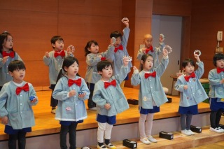 ステージに立つ水色のスモックと赤い蝶ネクタイ姿の十数名の園児たちが、カスタネットやすずを鳴らして演奏している発表会の写真