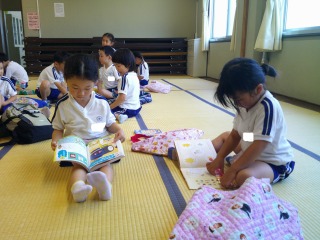 広い畳敷きの部屋で、体操服姿の複数の園児たちが座って思い思いの本を広げ、集中して本を見て過ごしている様子の写真