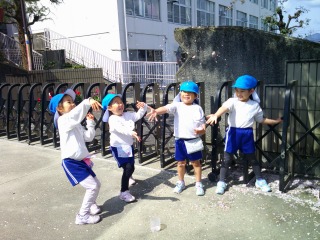 青い帽子と白い体操服姿の四人の園児が、黒いフェンスの前で満面の笑顔で踊っているような写真