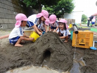 ピンク色の帽子をかぶった5人の園児が、砂場に作った大きな山のトンネルから水を流してとても楽しそうに遊んでいる写真