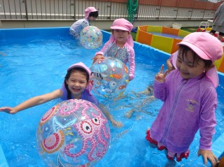 ピンク色の帽子とラッシュガードを着た四人の園児が、大きなボールで遊びながら、笑顔でビニールプールでの水遊びを楽しんでいる様子の写真