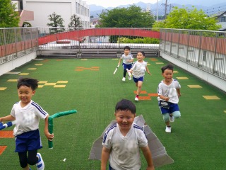 広々とした人工芝の園庭の上で、工作で作った翼やブーメランを持ち、五人の園児たちが満面の笑みで走っている写真