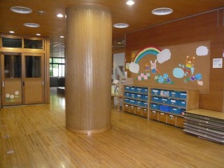 木目の床と壁に統一された明るい幼稚園の玄関ホールに、丸い柱と虹や動物のイラストが飾られた掲示板、そして園児のロッカーが設置されている室内の写真
