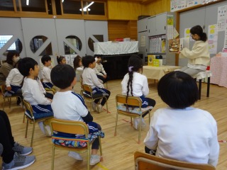 室内で、体操服姿の複数の園児たちが椅子に座って静かに、前にいる先生が読み聞かせする絵本に集中して聞いている写真