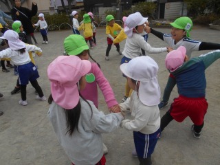 広場でピンクや黄緑など様々な色の帽子をかぶった複数の園児たちが手をつなぎ、楽しそうに輪になって遊んでいる様子の写真