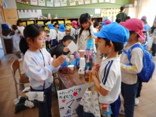 帽子をかぶった複数の園児たちが、積み重ねた各コップが置かれたテーブルの周りに並び、小学校のお姉ちゃんと向かい合って話をしている写真