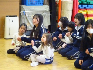 体操服を着た園児とジャージを着た女の子の生徒たちがフローリングの床に座って仲良く手をつないでいる写真