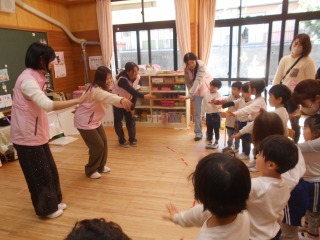 広々とした木の床の室内で、前で3名の先生が中腰になり右手を前に出しているのを複数の園児たちが真似をしている写真