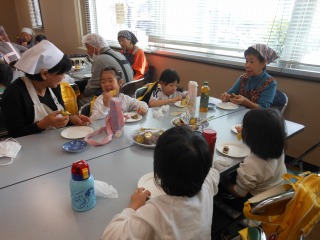 広いテーブルを囲み、園児たちが大人たちと交流しながら笑顔でサツマイモを美味しそうに食べている写真