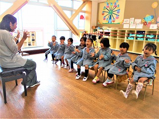 明るいフローリングの教室で、水色の制服を着た8人の園児たちが横一列に並べた椅子に座って、前に座った先生の手に合わせて真似をしている手遊びの写真