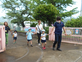 緑に囲まれたピンク色の門から、帽子とリュックを背負った三人の園児たちが保護者に手を引かれて登園している写真