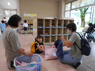 広々とした玄関ホールのロッカーの前で、保護者が荷物を置き、先生が籠の横で見守り、黄色いリュックを背負った園児が靴を脱いでいる登園時の写真