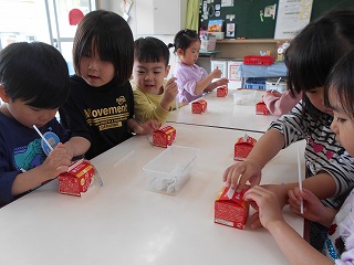 室内のテーブルで複数の園児たちが赤い小さな牛乳パックにストローを指している写真