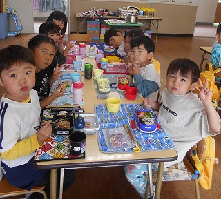 幼稚園の室内で8人の園児たちが二つの机を並べたテーブルを囲み、お弁当や水筒を広げて仲良く昼食をとっている写真