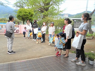 屋外の園舎前で、保育士が複数の保護者と園児に向かい合って連絡事項を伝えている様子の写真