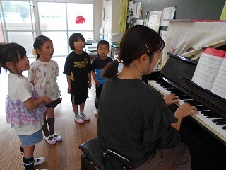 先生がピアノを弾き、その後ろに集まった四人の園児たちが、口を開けて歌を歌っている写真