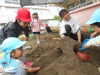 日陰のある屋外の砂場で、先生と砂山のトンネル作りに参加する赤と水色の帽子をかぶった複数の園児たちとのびのびと遊んでいる写真