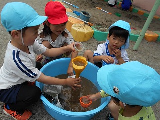 園庭の砂場の一角で、青や赤の帽子をかぶった四人の園児たちが大きな青いタライに溜めた泥水を、漏斗やカップを使って水遊びしている写真