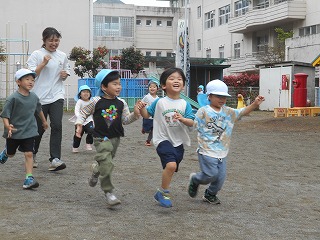 園庭で、青い帽子をかぶった複数の園児たちが先生と満面の笑顔で楽しそうに走り回っている写真