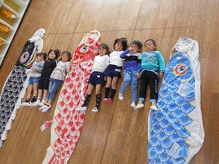 明るいフローリングの床の上で、7人の園児たちが大きな三匹の鯉のぼりの間で並んで寝そべっているのを上から撮影した写真