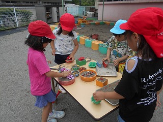 戸外のテーブルを囲む4人の園児が、赤い帽子や青い帽子をかぶり、砂を使って楽しそうに四角や丸の型枠に砂を入れ色とりどりの小石や花びら飾った土遊びの写真