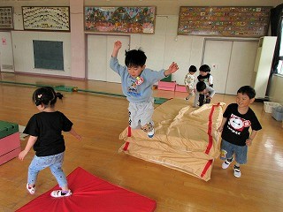 屋内で数名の園児たちが、積み重ねたマットの上を跳び箱のように飛び越えたり、赤いマットの上を歩いたりして遊んでいる様子の写真