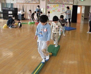 広い室内で園児たちが、緑色の細長い平均台の上をバランスを取りながら慎重に歩いたり、カラフルなステップ台の上で運動している様子の写真