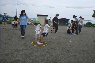 園庭で、マスクを着用した先生や保護者が見守る中、複数の園児たちがフラフープを使ったゲームや手をつないで歩くなどの集団遊びをしている様子の写真