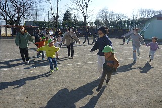 よく晴れた日の屋外で、園児たちとマスクを着用した先生や保護者と手をつなぎながら広場を駆け回って遊んでいる楽しげな様子の写真