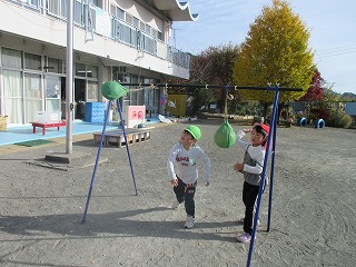 屋外の園庭で、二人の園児が吊るされた緑色の丸い玉を目がけて勢いよくパンチをしてあ遊んでいる様子の写真