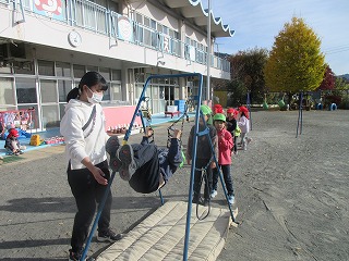 マスクを着用した女性の先生が遊具につかまって逆上がりをする園児を補助し、後ろには他の園児たちが順番を待って並んでいる様子の写真