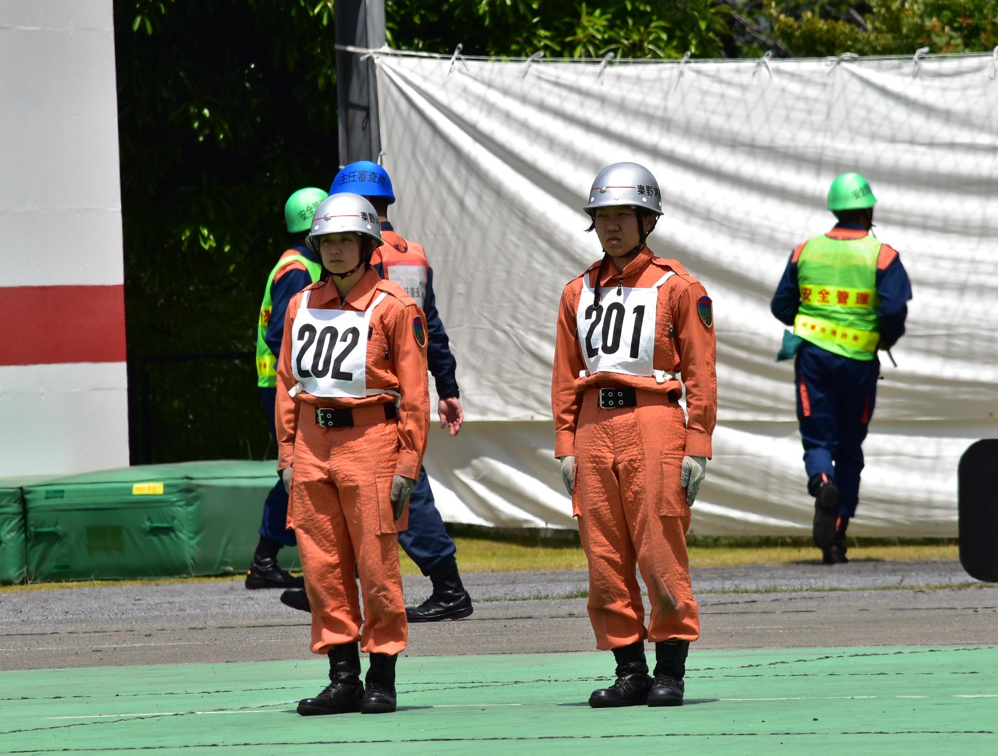 オレンジ色のレスキュー服に番号「202」と「201」のゼッケンをつけ、銀色のヘルメットをかぶった2名の女性隊員が、緑色に塗られた地面の上に立ち、背後には他の隊員や白い幕が見える、訓練中の写真