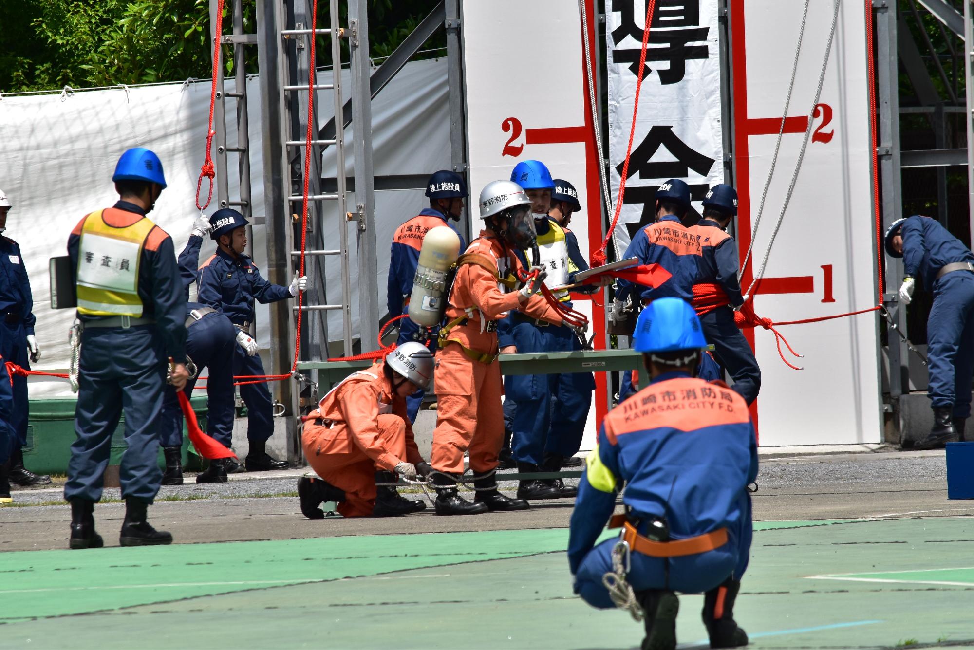 酸素ボンベを背負った女性隊員や濃紺とオレンジ色の活動服を着た複数の消防隊員が、資機材を使用してロープを用いた救助訓練に取り組んでいる様子の写真