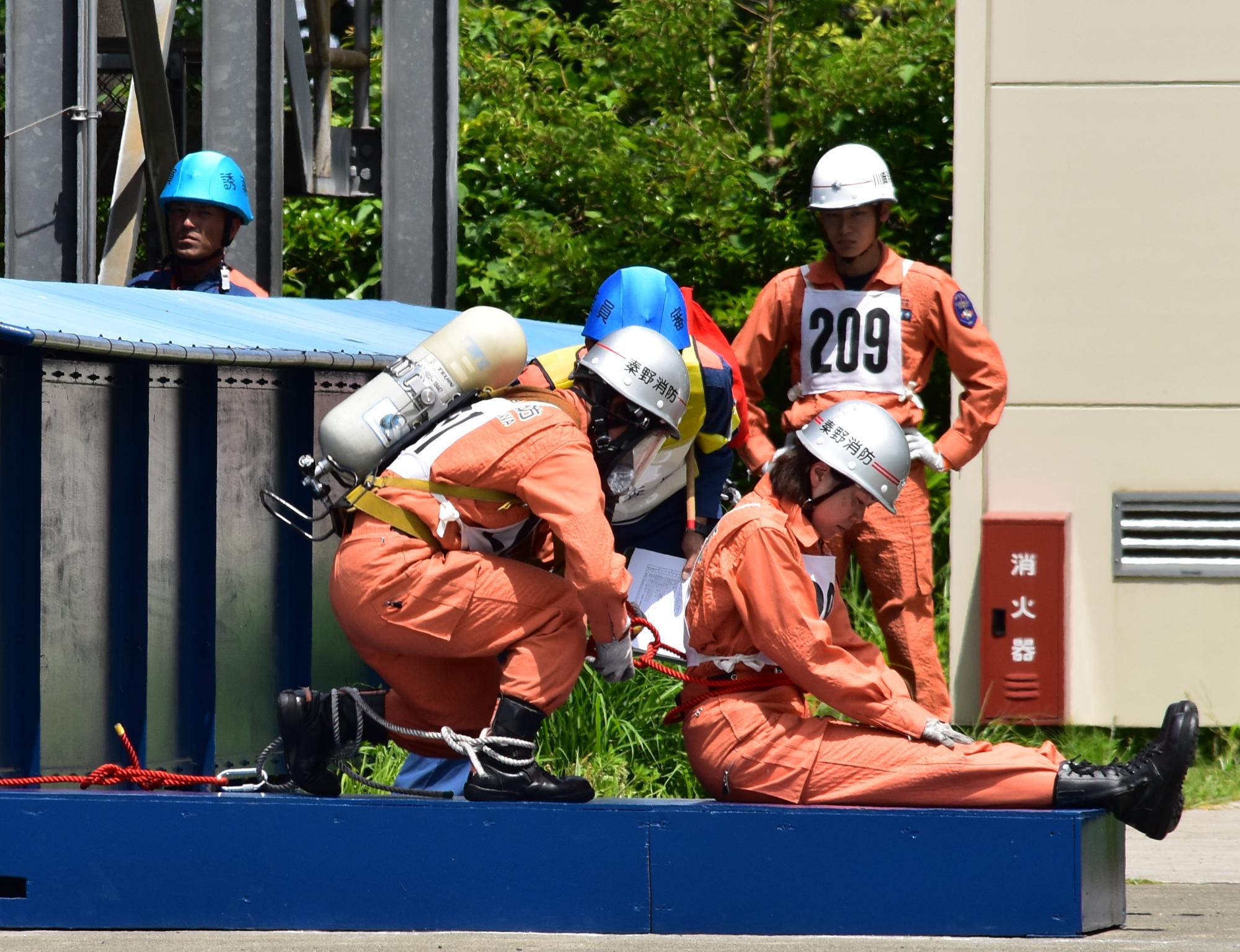 番号「209」のオレンジ色のレスキュー服を着た隊員が立っている手前で、酸素ボンベを背負った隊員が、青い台の上に座った別の隊員の腰にロープを結んでいる救助訓練の様子の写真