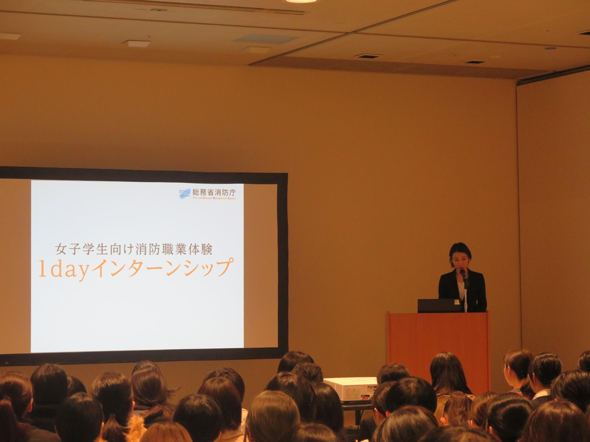 ホールで、女性がマイクを使ってプレゼンテーションをしており、その後ろのスクリーンには「女子学生向け消防職業体験1dayインターンシップ」の文字が映し出されている写真