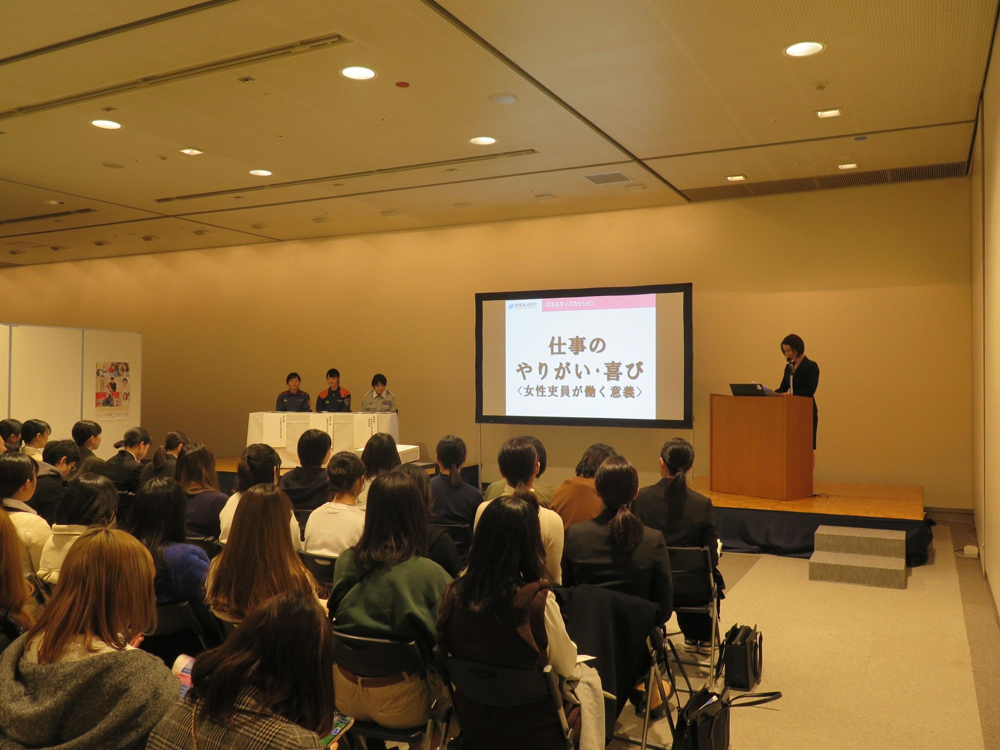大勢の参加者が着席したホールで、「仕事のやりがい・喜び」と表示された壇上のスクリーンの左側の席に、3人の消防職員が座っている様子を写した写真