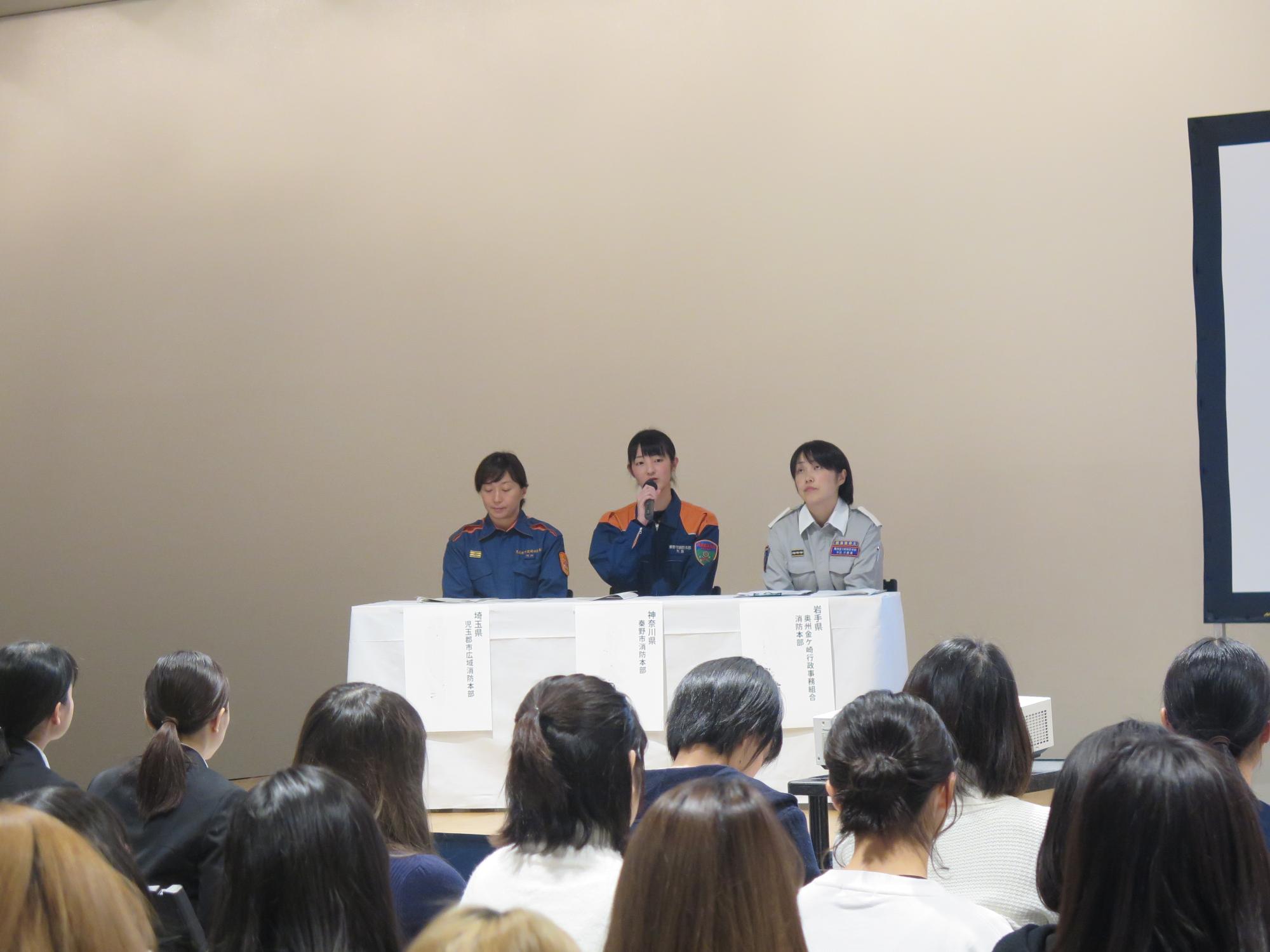 多数の聴衆が座っている前で、三人の女性消防吏員が壇上のテーブルに座り、真ん中の女性がマイクを持って話している写真