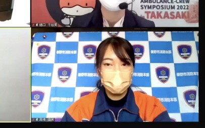 レスキュー服姿で薄い茶色のマスクを着用した女性が、秦野市消防本部のロゴと文字の入った青と白のパネルを背景にして、オンライン会議に参加している様子の画面キャプチャ画像