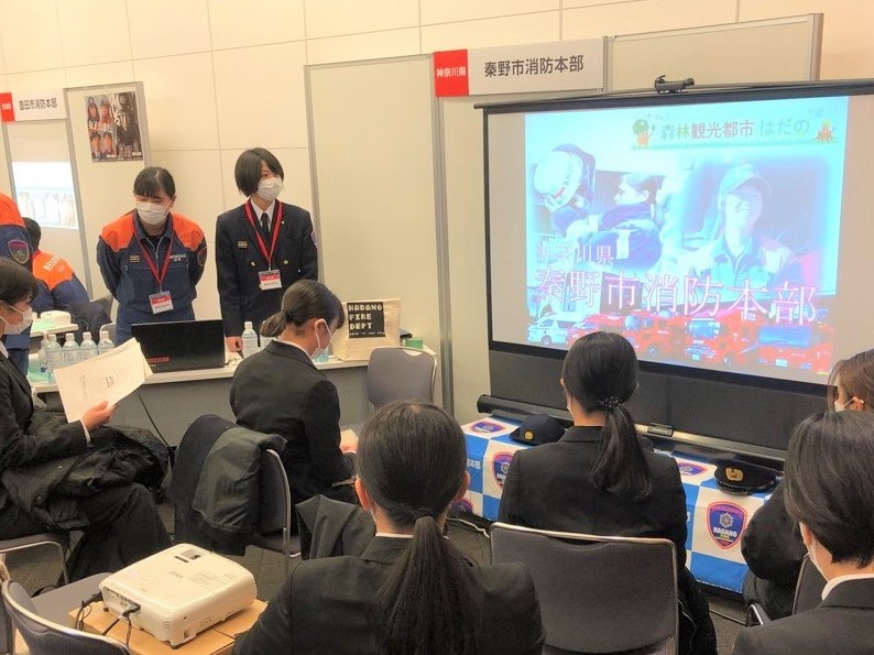 制服や活動服姿の女性職員が、座っている複数の学生たちに向けて、「秦野市消防本部」の文字が映し出されたスクリーンを使って説明をしている様子の写真