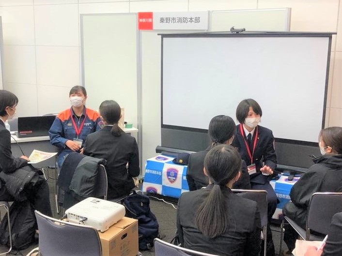 濃紺の制服を着用した女性職員とオレンジ色の活動服の女性職員の計2名が、席に座った学生たちに、対話形式で説明を行っている様子の写真