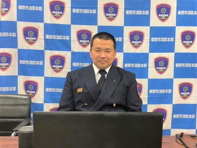 秦野市消防本部のロゴと文字が格子状に印刷された青と白のパネルを背景に、濃紺の制服を着用した男性職員がノートパソコンの前に座り、カメラに向かって真剣な表情を浮かべている様子の写真